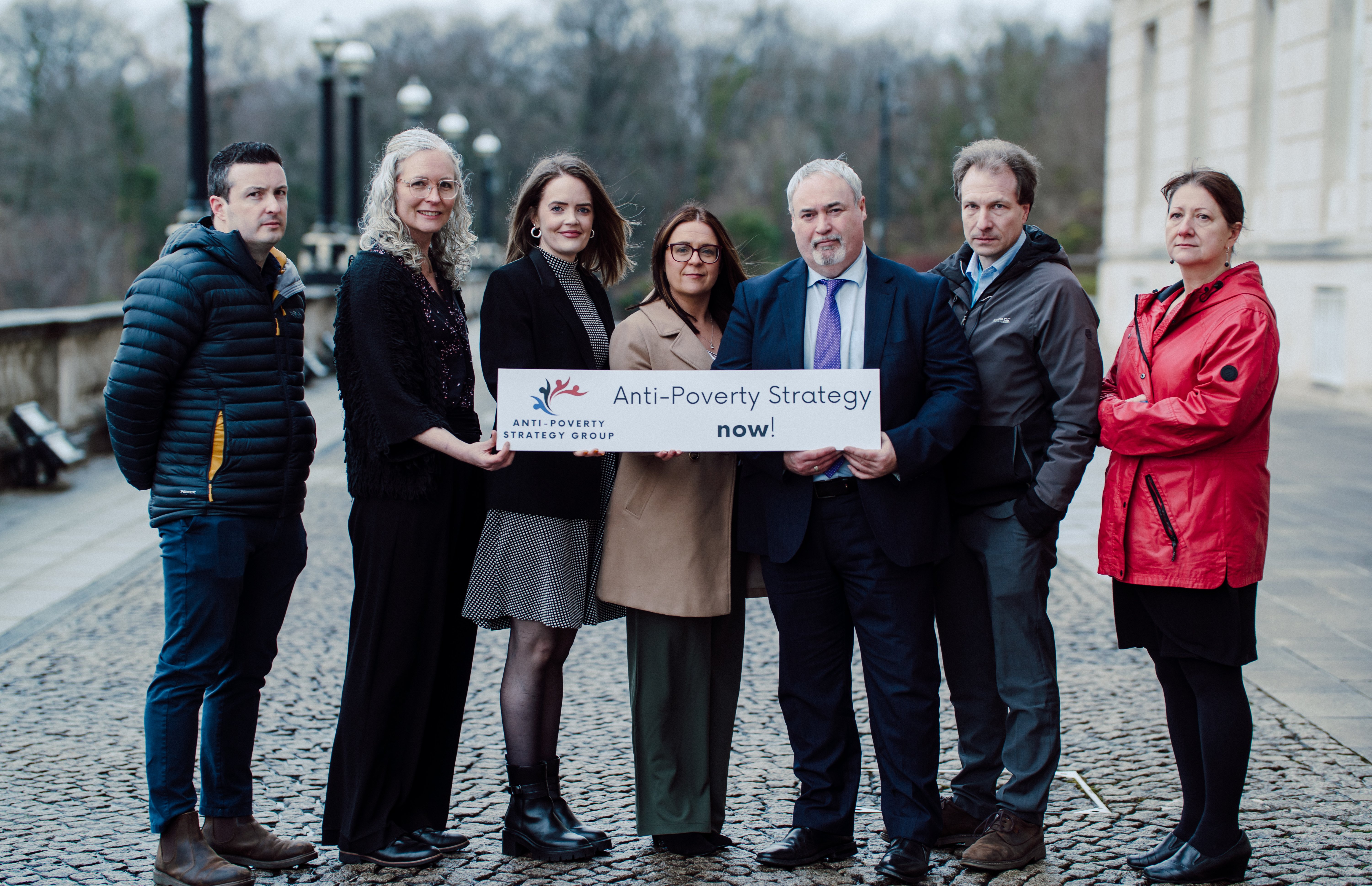 group outside Stormont holding "anti-poverty strategy now!" sign