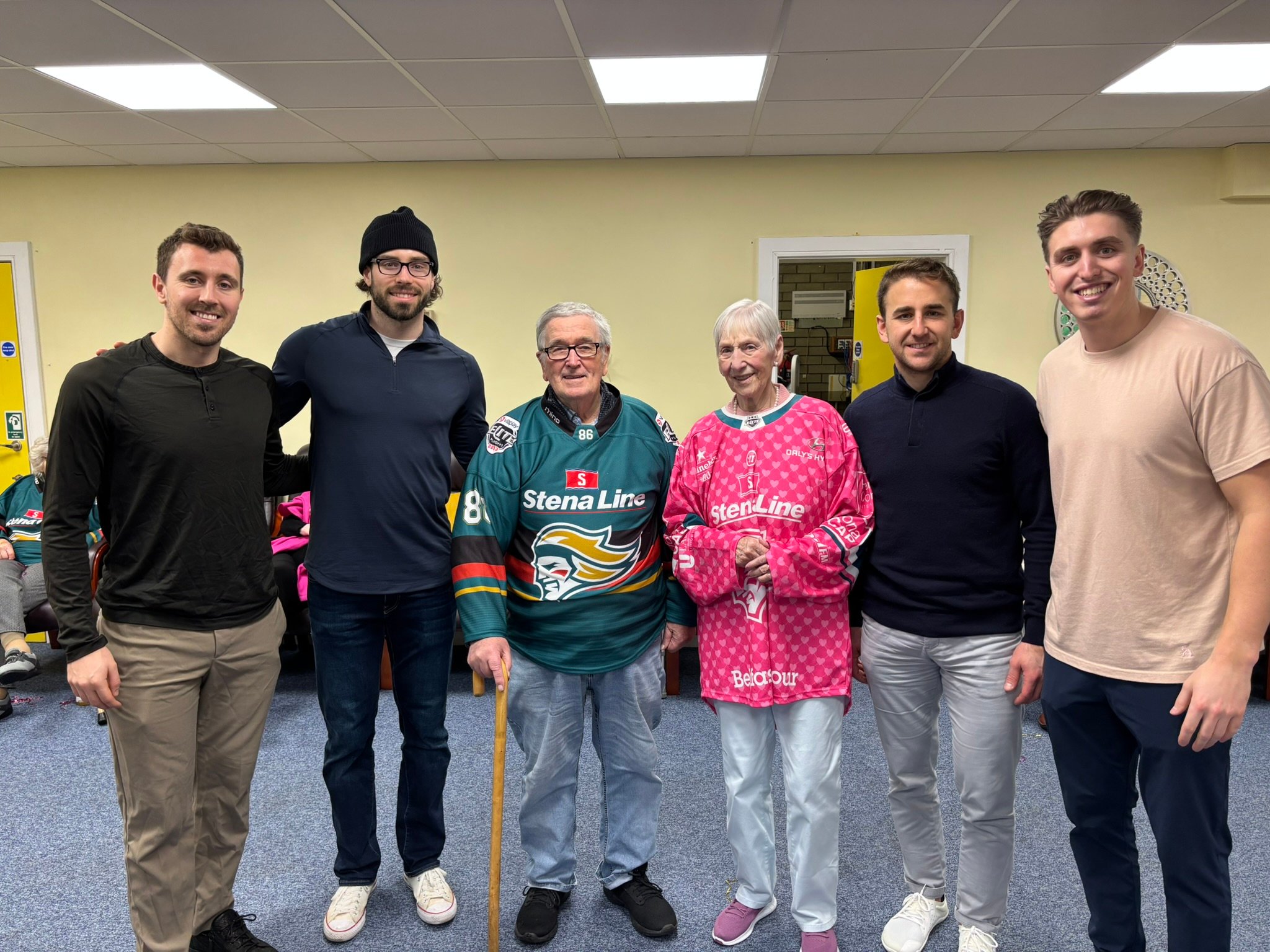 four Belfast Giants ice hockey players standing on either side of two older people wearing Giants jerseys